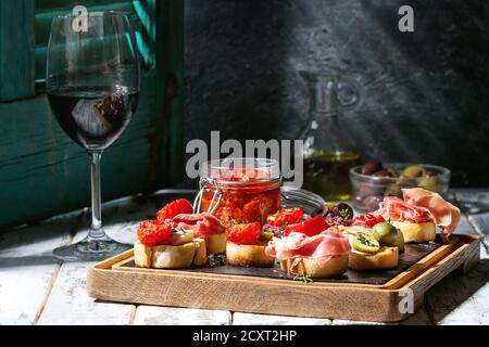 Tapas or bruschetta variety. Bread with ham prosciutto, sun dried tomatoes, olive oil, olives, pepper on slate wood board with glass of red wine over Stock Photo