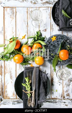 Christmas table decorations with clementines or tangerines with leaves and green branches on black ornate board. Empty plate with cutlery and textile Stock Photo