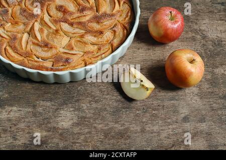 Traditional french homemade apple pie in a white ceramic baking form and autumn apples on old dark wooden background. Copy space Stock Photo