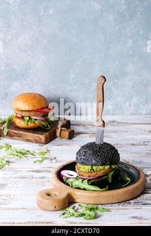 Set of homemade burgers in black and white buns with avocado, tomato sauce, arugula, cheese, onion on wood serving board over white wooden plank table Stock Photo