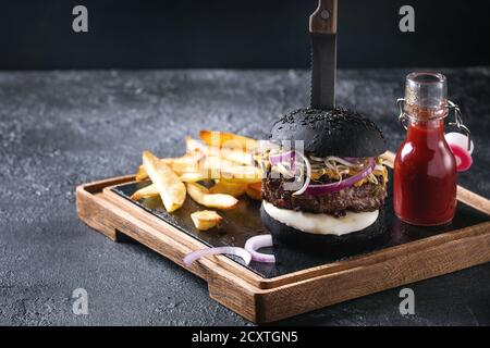 Homemade black bun hamburger with beef, mozzarella cheese, sprouts served on wooden slate serving board with french fries, knife and ketchup sauce on Stock Photo