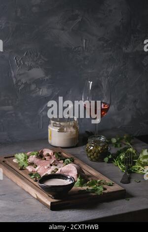 Vitello tonnato italian dish. Thin sliced veal with tuna sauce, capers and coriander served on wooden serving board, glass of rose wine and ingredient Stock Photo