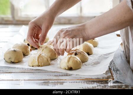 Raw unbaked buns. Ready to bake homemade Easter traditional hot cross buns on baking paper over white wooden table. Window at background. Female hands Stock Photo