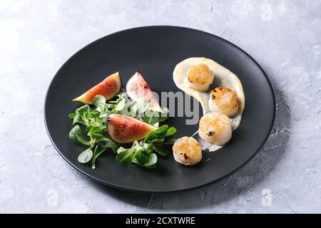 Fried scallops with lemon, figs, sauce and green salad served on black plate over gray texture background. Plating, fine dining Stock Photo