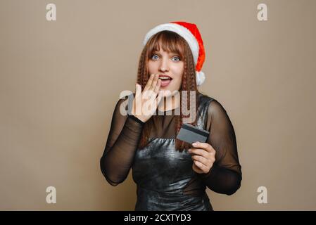Young woman in Santa hat, demonstrates blank credit card over beige wall, Christmas shopping time begins. Christmas holiday, Banking Business, Shopping concept. Stock Photo