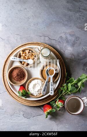 Ingredients for cooking chocolate mug cakes. Flour, cocoa powder, sugar, caramel in wooden bowls, milk, strawberries and mint served with spoons and m Stock Photo