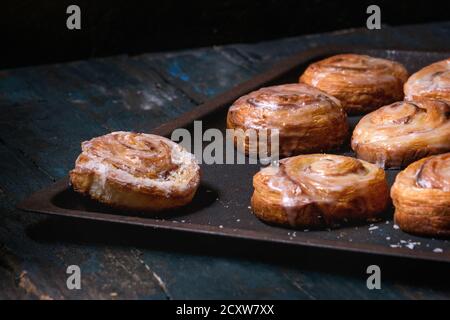 Homemade glazed puff pastry cinnamon rolls with custard and raisins on oven tray over old dark blue wooden background. Rustic style Stock Photo