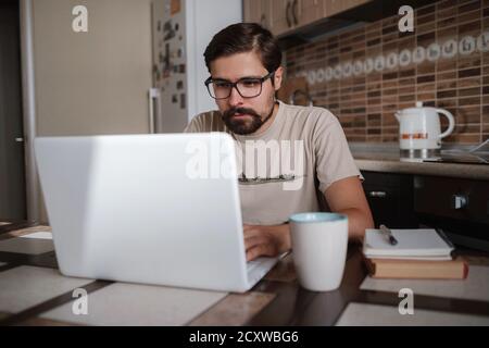 Focused young man wearing glasses using laptop, typing on keyboard, writing email or message, chatting, shopping, successful freelancer working online Stock Photo