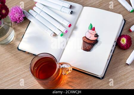 sketching cupcake in a notebook. All placed on a wooden table with a transparent cup of tea, notebook, markers, and flowers. Top view Stock Photo