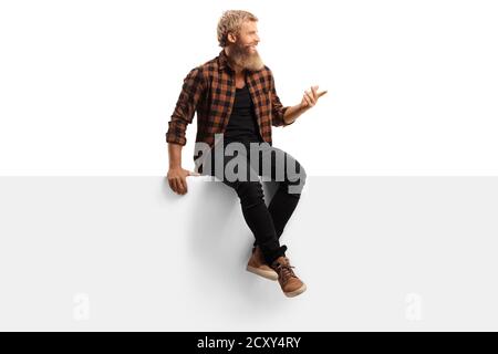 Young bearded man sitting on a white panel board and gesturing with hand isolated on white background Stock Photo
