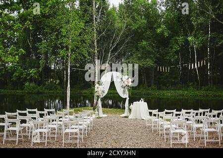 Outdoors wedding ceremony set. Wedding arch decorated with fresh flowers and empty chairs placed near the small lake on the open air. Stock Photo