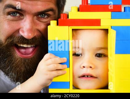 Boy and man play together, close up. Father and son with happy and serious faces hold colorful toy bricks construction. Toys and childhood concept. Dad and kid hide behind house made of plastic blocks Stock Photo