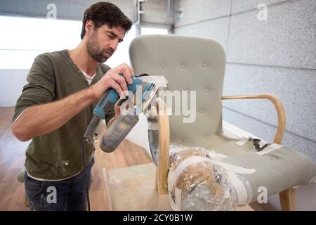 A white man power sanding the arm of an antique chair Stock Photo