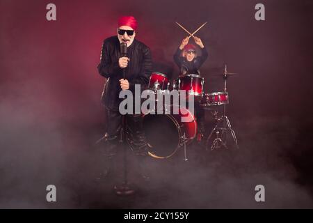 Photo of pensioners lady man popular rock group couple play drum instruments sing famous song concert show wear rocker leather outfit bandana sun Stock Photo