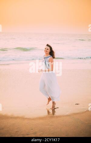 Goa, India. Young Caucasian Woman In White Dress Walking Along Seashore, Looking At Camera And Smiling In Summer Sunlight Stock Photo