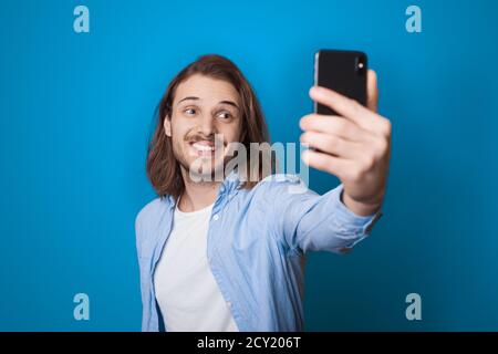 Young long haired man with beard is smiling and making a selfie using a phone on a blue studio wall in casual clothes Stock Photo