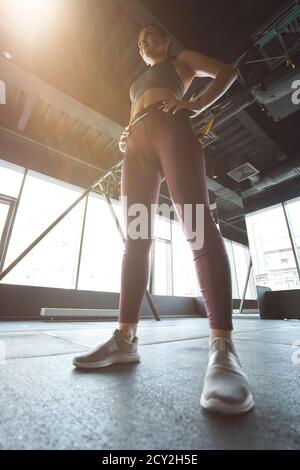 Bottom view of a strong athletic woman in sportswear keeping arms on hips while working out at industrial gym, vertical shot. Sport, wellness and healthy lifestyle Stock Photo