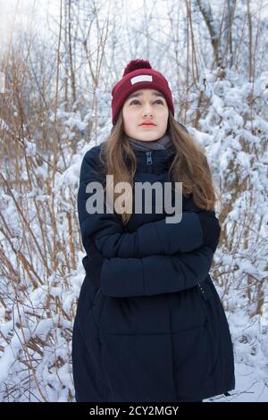 Beautiful girl freezing in winter forest. Portrait of serious cute attractive young teen girl with long blonde hair with green eyes on natural blurred Stock Photo