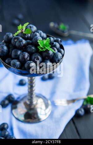 Top view blueberries in vintage metal bowl decorated with mint leaves on the blue napkin on a black wooden background. Dark mood photo. Vertical card Stock Photo