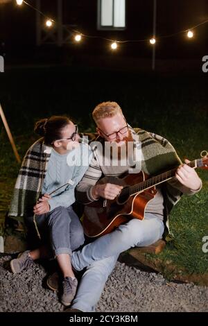 Young couple, caucasian woman and red haired bearded man, on a night picnic with a guitar and marshmello have a good time Stock Photo
