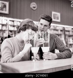 1950s DATING TEENAGE COUPLE BOY WATCHING GIRLFRIEND DRINKING SODAS PAPER CUP PAPER STRAW AT DRUG STORE LUNCH COUNTER FOUNTAIN - f8341 HAR001 HARS PAIR ROMANCE BEAUTY SUBURBAN FOUNTAIN URBAN SHARING STRAW PEARL OLD TIME NOSTALGIA OLD FASHION 1 JUVENILE STYLE COMMUNICATION BALANCE TEAMWORK JOY LIFESTYLE FEMALES HEALTHINESS SEATED COPY SPACE FRIENDSHIP HALF-LENGTH PERSONS BOYFRIEND CARING MALES TEENAGE GIRL TEENAGE BOY DRUG B&W DATING GIRLFRIEND TEMPTATION PRETTY HAPPINESS WELLNESS LEISURE STRATEGY CHOICE RECREATION AT OPPORTUNITY SODAS CONNECTION CONCEPTUAL STYLISH TEENAGED INFATUATED GROWTH Stock Photo