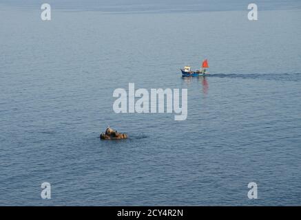 Working fishing boat heading for lobster pots passing rocky outcrop Stock Photo