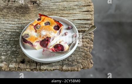 A slice of homemade clafoutis cherry pie - traditional french dessert in a gray plate and a spoon on the old wooden table close-up Stock Photo