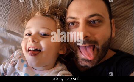 Authentic close up of a dad and daughter making funny faces, looking at the screen for a selfie, in a bed. Concept of family and emotional relationshi Stock Photo