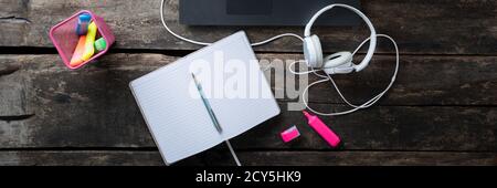 Wide view image of open blank notepad lying on rustic wooden desk by a laptop computer  with headphones and colorful markers next to it. Stock Photo
