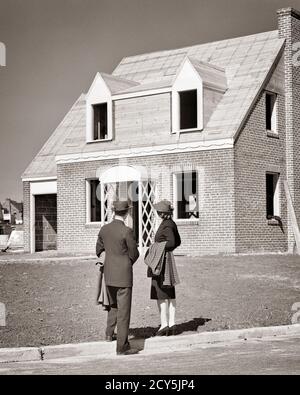 1930s 1940s YOUNG COUPLE MAN WOMAN HUSBAND WIFE BACK VIEW LOOKING AT NEW HOME UNDER CONSTRUCTION IN HOUSING DEVELOPMENT  - s5180 HAR001 HARS FEMALES MARRIED SPOUSE HUSBANDS HOME LIFE COPY SPACE FULL-LENGTH LADIES PERSONS MALES BUILDINGS B&W PARTNER DEVELOPMENT DREAMS PROPERTY YOUNG MAN EXTERIOR AT IN PURCHASE BUYERS REAL ESTATE STRUCTURES INVESTMENT EDIFICE MID-ADULT MID-ADULT MAN MID-ADULT WOMAN PROSPECTIVE TOGETHERNESS TWO STORY WIVES BLACK AND WHITE CAUCASIAN ETHNICITY HAR001 OLD FASHIONED Stock Photo
