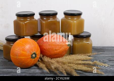 Jam in jars. Nearby is a pumpkin and a bouquet of dry grass with spikelets. On black pine boards. Stock Photo