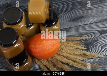 Jam in jars. Nearby is a pumpkin and a bouquet of dry grass with spikelets. On black pine boards. Stock Photo