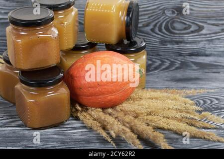 Jam in jars. Nearby is a pumpkin and a bouquet of dry grass with spikelets. On black pine boards. Stock Photo