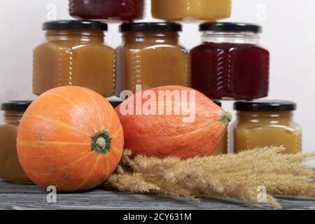 Jam in jars. Nearby is a pumpkin and a bouquet of dry grass with spikelets. On black pine boards. Stock Photo