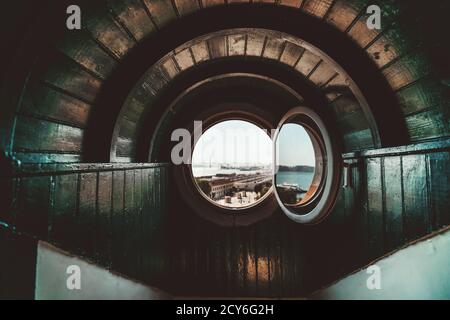 A wide-angle view of a wooden arc in a nook recess of a house with a round opened window or a porthole with a daytime cityscape in a defocused backgro Stock Photo