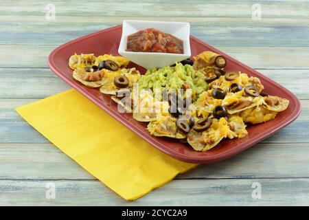 Nachos with cheese, bean dip, and clack olives with guacamole in middle and with salsa in small white dipping bowl on red platter with yellow napkin Stock Photo