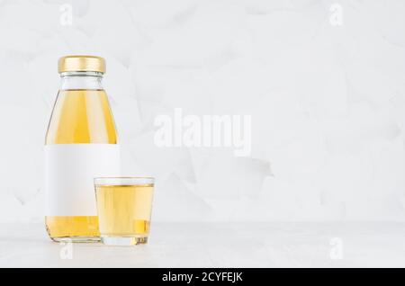 Fresh yellow transperent apple juice in glass bottle mock up with blank label,  wine glass on white wood table in light interior, template for packagi Stock Photo