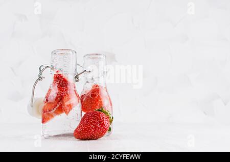 Refreshing cold fruit drinks with red strawberry, bright bubbles  in transparent yoke bottles in elegant white interior on wood table, copy space. Stock Photo