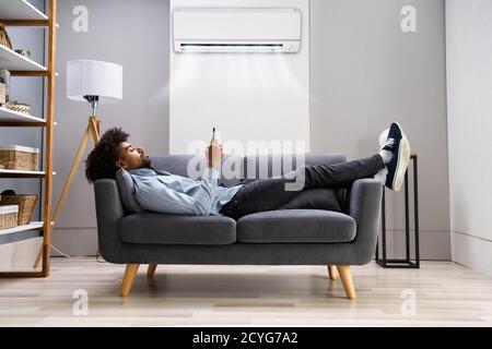 Young Man Lying Under Air Conditioner On Couch Using Smartphone At Home Stock Photo