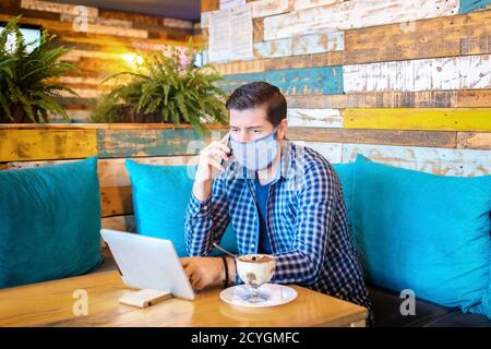 Relaxed entrepreneur with protective face mask remote working at digital tablet while talking on mobile phone in coffee shop Stock Photo