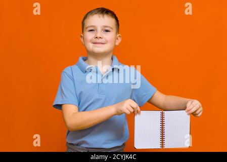 Cuccessful caucasian boy in a blue t-shirt holds empty notebook paper advertising.Boy shows to the your banner.Orange studio wall. Stock Photo