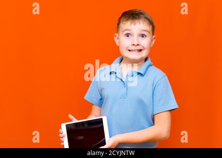 Portrait of european schoolboy shows tablet and blue t-shirt on orange studio background.cheerfully thinks something and showing something on empty ta Stock Photo
