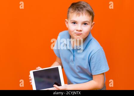 Portrait of european schoolboy shows tablet and blue t-shirt on orange studio background.cheerfully thinks something and showing something on empty ta Stock Photo