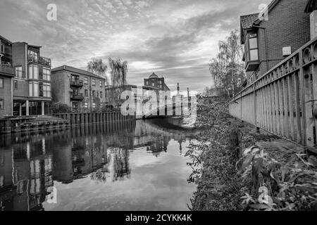 Black and white photo of a view down the River Wensum in the city of Norwich, Norfolk. Captured on the riverside footpath in the residential area looking towards the historical White Friar's Bridge. Stock Photo