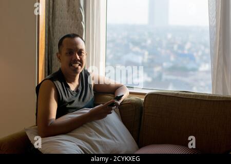 Asian man in armless clothes sitting in room near clear glass window, eyes looking at smart phone and using mobile phone in hand holding Stock Photo