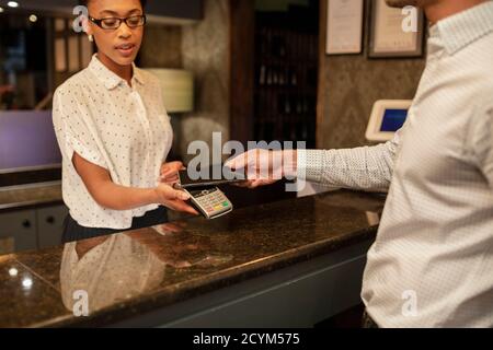 A female hotel receptionist taking the contactless payment of a customer who is checking in to the hotel, he is using his smart phone to pay. Stock Photo