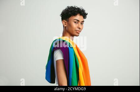 Handsome young man with pride movement LGBT Rainbow flag on shoulder against white background. Man with a gay pride flag looking at camera. Stock Photo