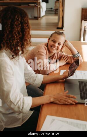 Woman talking with her daughter while working at home. Girl smiling and listening to her mother holding digital tablet. Stock Photo