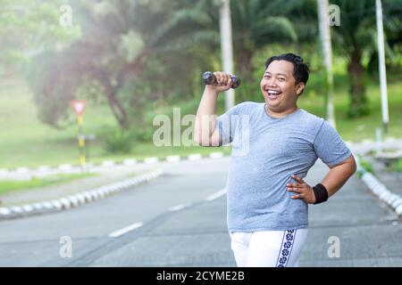 Asian young man with obesity confidently lifts a barbell and poses in front of the camera in an outdoor city park Stock Photo
