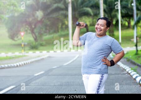 Asian young man with obesity confidently lifts a barbell and poses in front of the camera in an outdoor city park Stock Photo